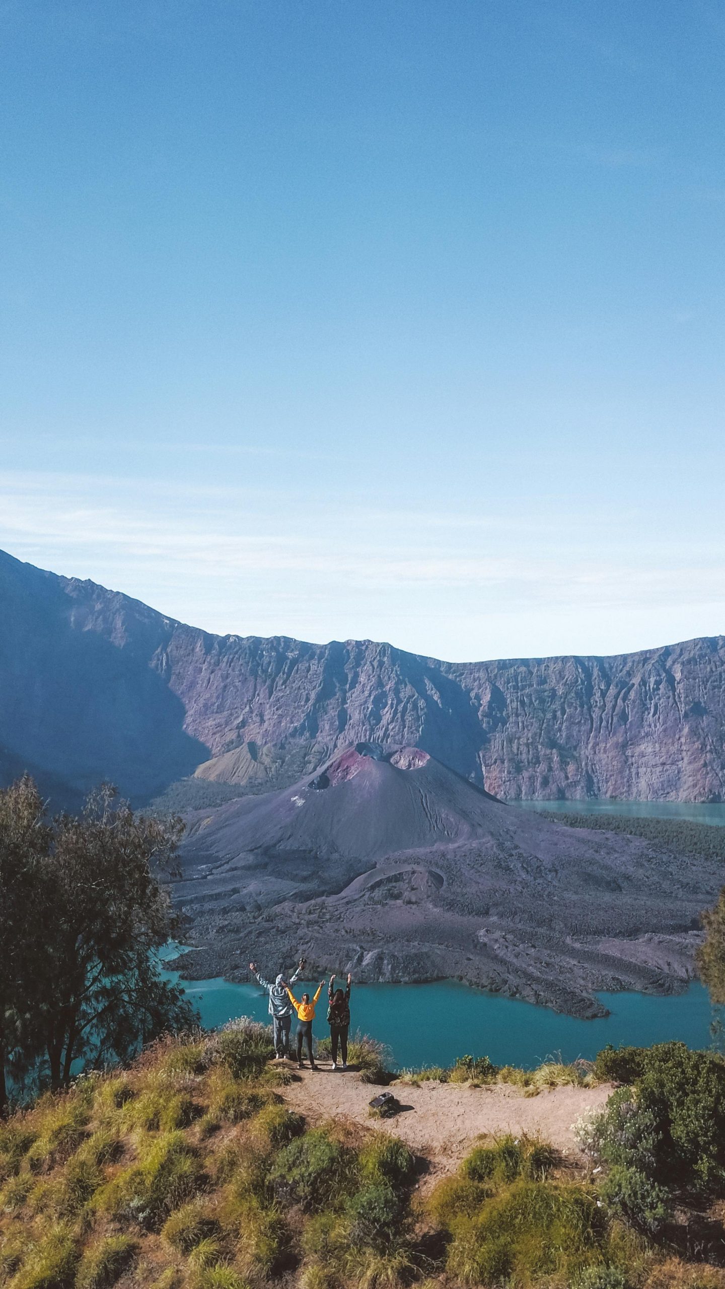 Hikers raising arms in triumph on Mount Rinjani with a volcanic backdrop and clear sky.